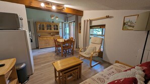 Ein gemütliches Wohnzimmer mit einem Holztisch, einem Sessel und einer Couch. Eine Küchenzeile mit Holzausbau und einem Kühlschrank ist ebenfalls vorhanden.