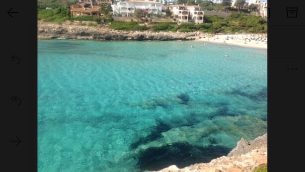 A coastal area with clear turquoise water, rocky shoreline, and buildings in the background. - PENTHOUSE APARTMENT CALA ANGUILA BALCONY (Cala Anguila)
