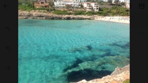 A coastal area with clear turquoise water, rocky shoreline, and buildings in the background.