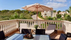 A balcony with a table, chairs, and an umbrella. There is a view of a pathway leading to a building with a white facade. - PENTHOUSE APARTMENT CALA ANGUILA BALCONY (Cala Anguila)