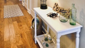 Un pasillo con piso de madera, un banco blanco con lámpara, una mesita con objetos decorativos y una puerta al final.