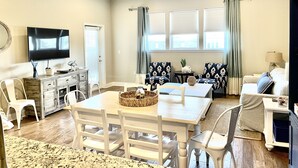 Un salón amplio con ventilador de techo, televisor de pantalla plana, mesa de comedor con sillas y una cama para mascotas.