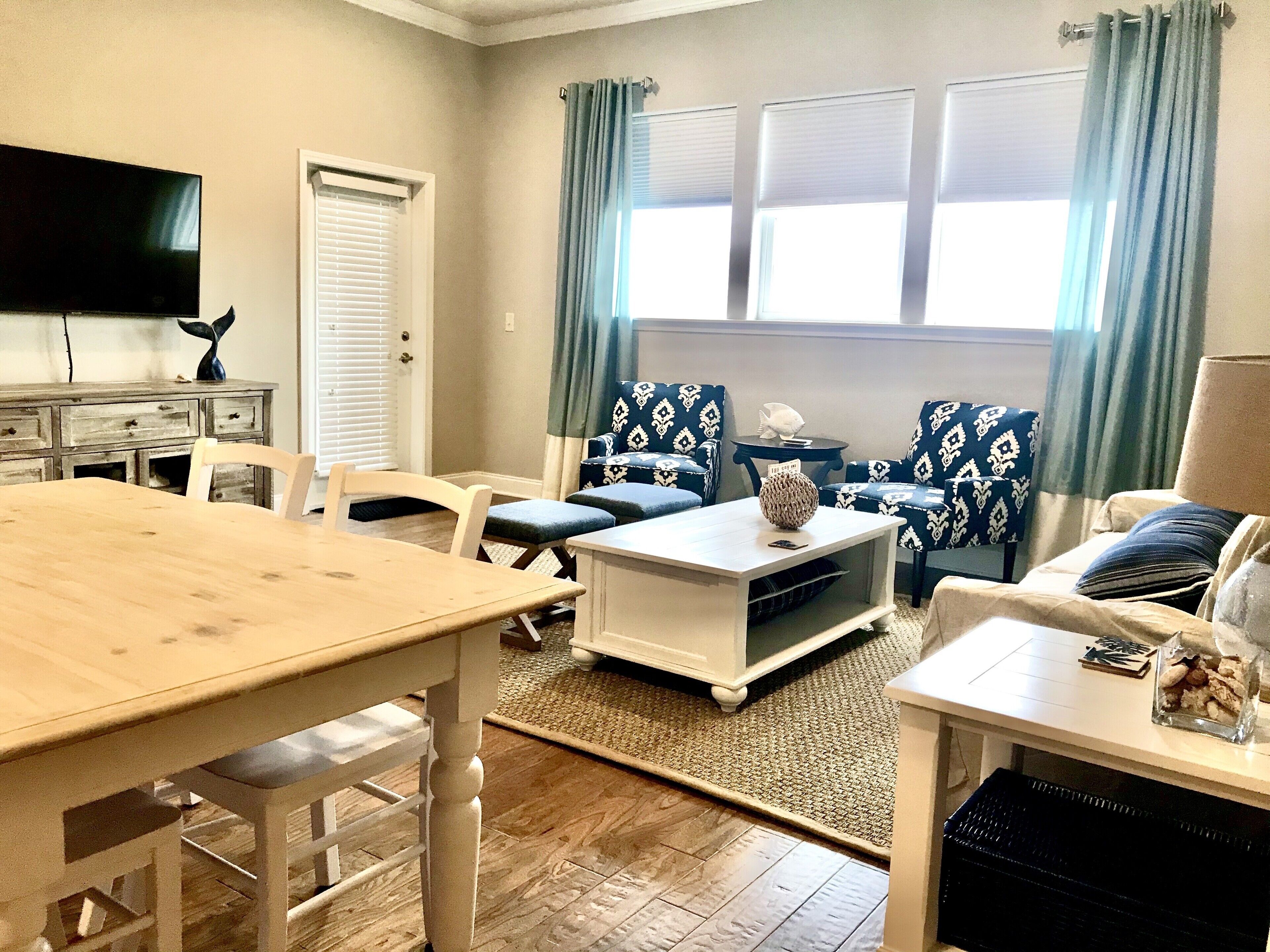 Una sala de estar con una mesa de madera, dos sillones azules estampados, una mesa baja blanca y un televisor en la pared.