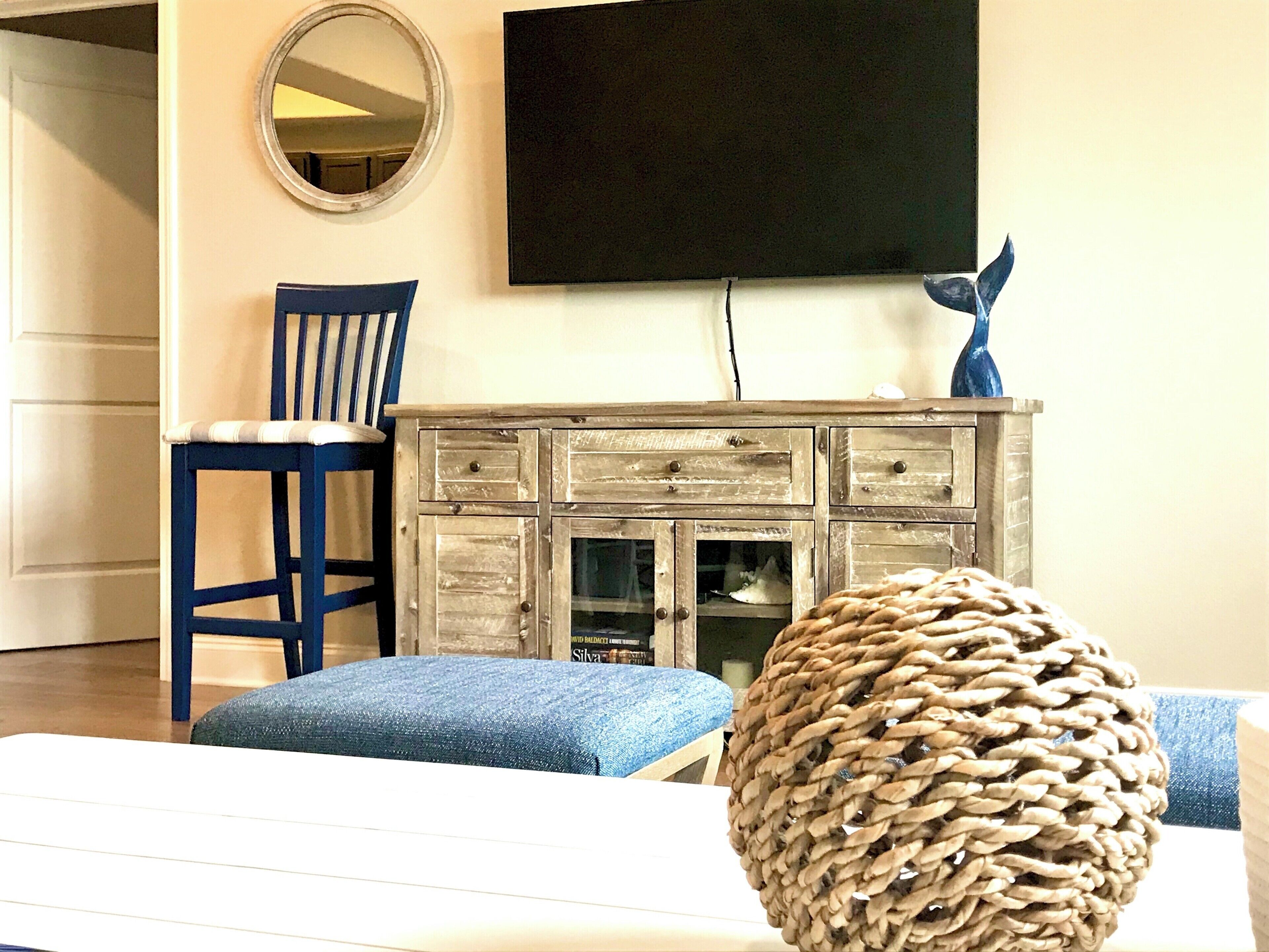 Ein Wohnzimmer mit einem an der Wand befestigten Fernseher, einem Holzkabinett, einem blauen Sessel und einem gewebten Pouf.