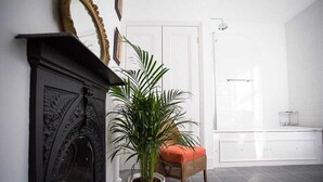 Ein Zimmer mit Kamin, einer Topfpflanze, einem Korbstuhl mit orangefarbenem Kissen und einer Glasdusche.