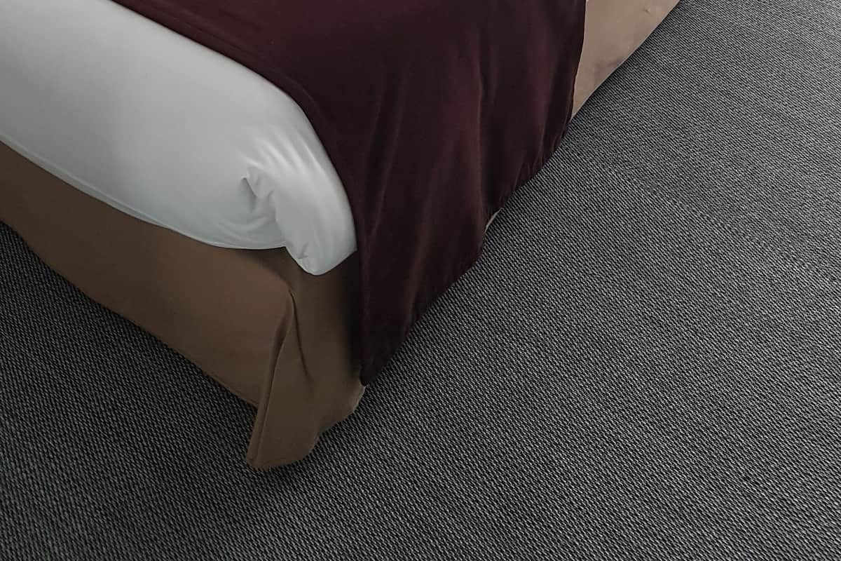 Una cama bien hecha con sábanas blancas y una manta marrón, cabecera oscura y una mesita de noche con lámpara.