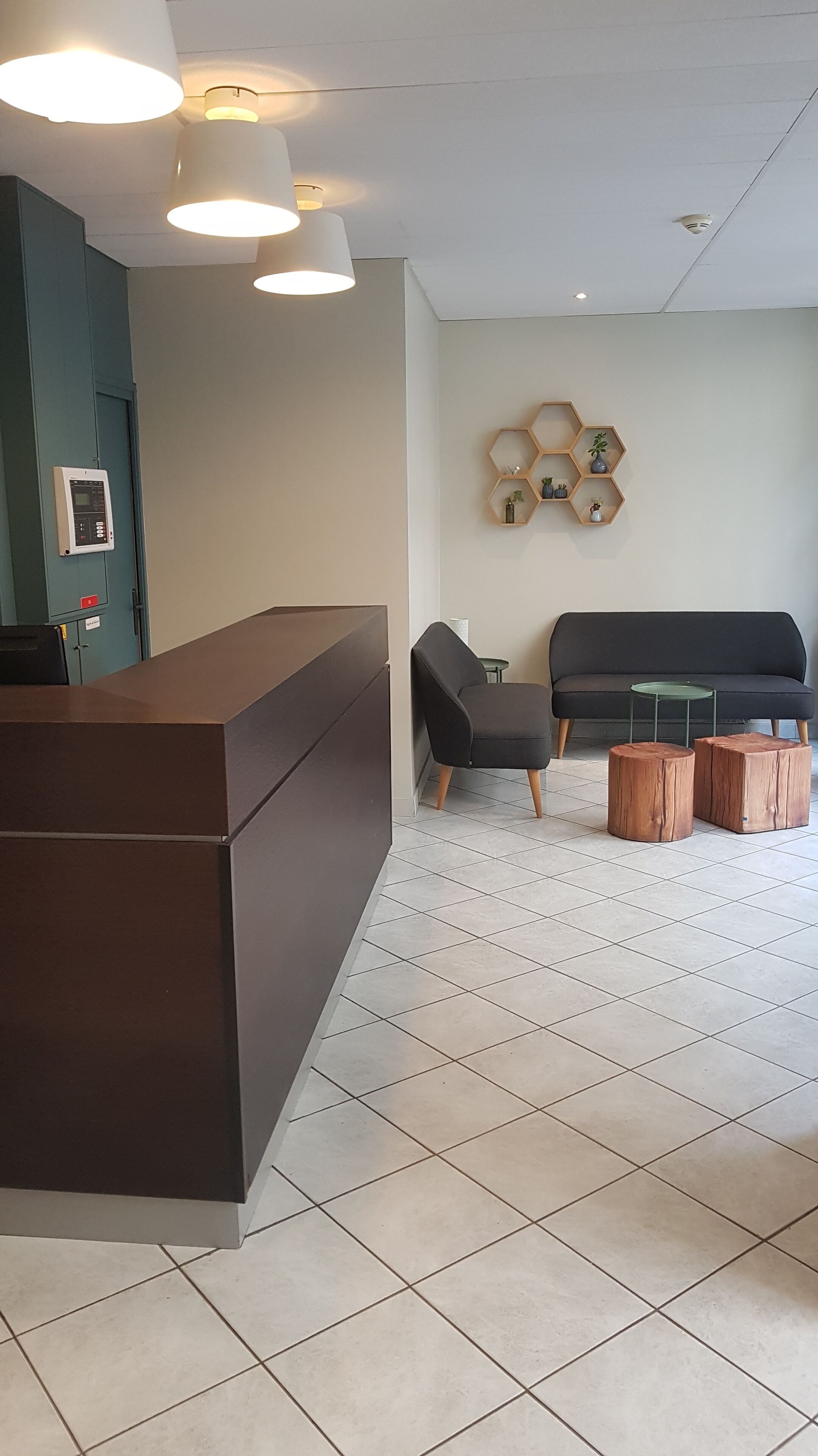 A reception area with a dark reception desk, a seating area with two chairs and a small table, and a wall-mounted hexagonal shelf with decorative items.