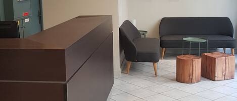 A reception area with a dark reception desk, a seating area with two chairs and a small table, and a wall-mounted hexagonal shelf with decorative items.