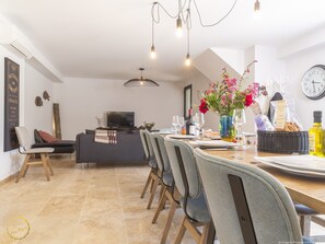 A modern dining area with a long table set for six, a television in the background, and a decorative vase with flowers.