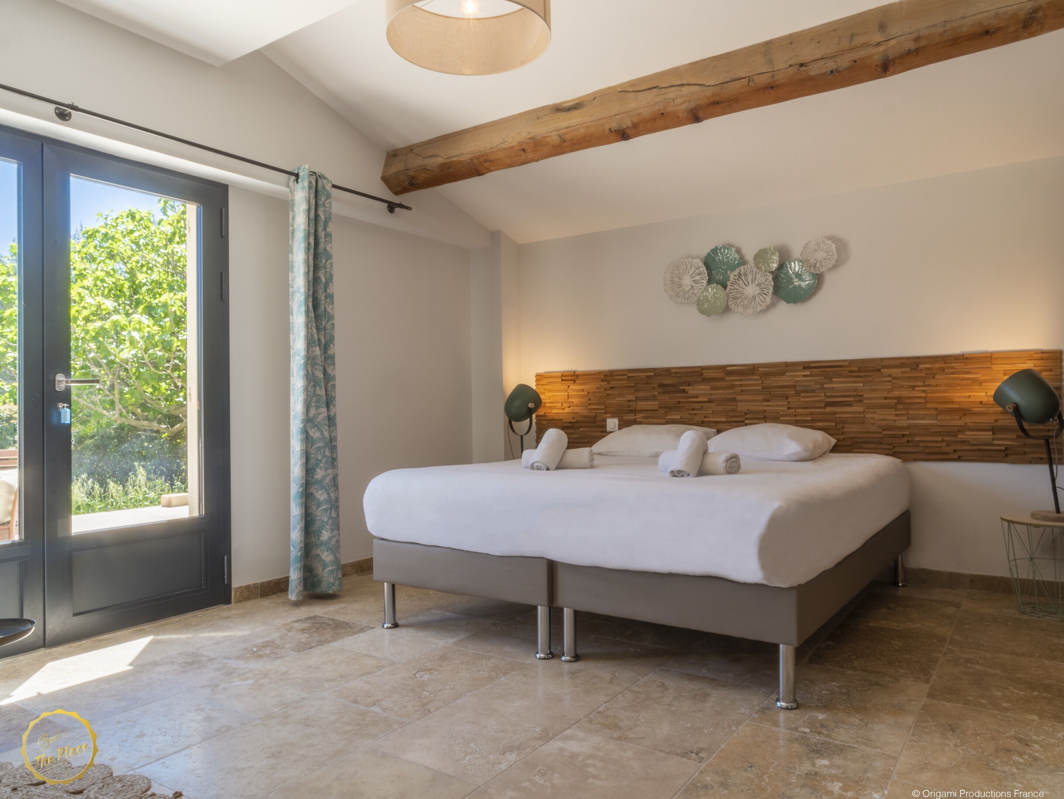 A modern bedroom with a large bed, a wooden ceiling beam, a glass door leading to a balcony, and a wall-mounted lamp.
