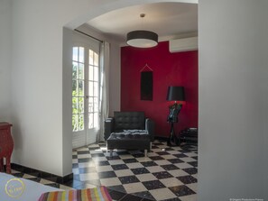 A room with a checkered floor, a red accent wall, a black armchair, a lamp, and a window with curtains.