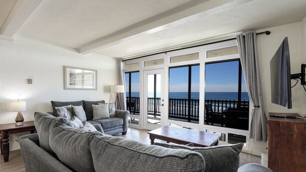 A living room with a grey sofa, a coffee table, and a TV on a wooden stand. There is a large window with a view of the sea.