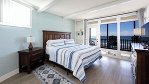 A bedroom with a large bed, a nightstand, a dresser, a window, and a view of the ocean through a sliding glass door.