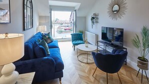 A modern living room with a blue sofa, a teal armchair, a round coffee table, and a TV. - Host & Stay | The Penthouse at Prospect Hill Apartments (Whitby)