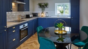 A modern kitchen with dark blue cabinets, a glass dining table, teal chairs, and a window with a view of greenery. - Host & Stay | The Penthouse at Prospect Hill Apartments (Whitby)