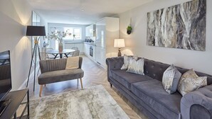 A modern living room with a grey sofa, a coffee table, and a floor lamp. There is a dining area with a table and chairs in the background.