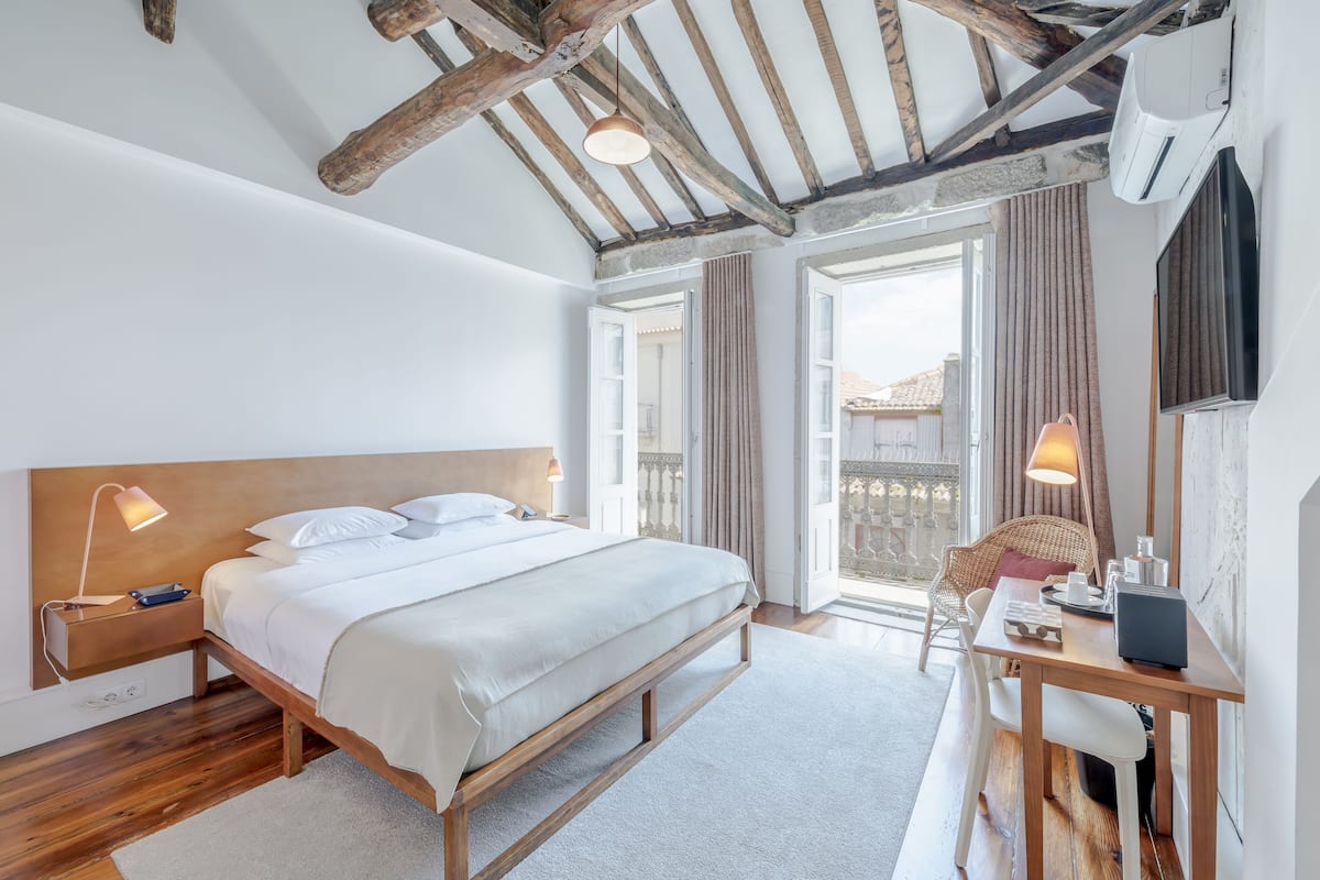 Un dormitorio con una cama de madera, lámparas de noche, un escritorio con silla y un balcón con cortinas.