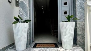 A building entrance with a marble facade, a nameplate reading 'BUILDING JOHN PASCHALIS,' and two large potted plants. - Grecian Sea (Thasos)