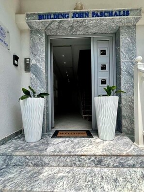 Entrada de un edificio con fachada de mármol, una placa que dice 'BUILDING JOHN PASCHALIS' y dos grandes macetas con plantas.
