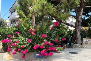 Um edifício com varanda, rodeado por vegetação e flores cor-de-rosa.