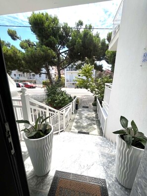 Uma varanda com plantas em vasos, grade branca e vista para a rua e árvores.