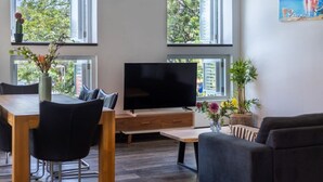Una sala de estar moderna con un televisor de pantalla plana, muebles de madera y ventanas grandes que ofrecen vista a árboles y edificios.