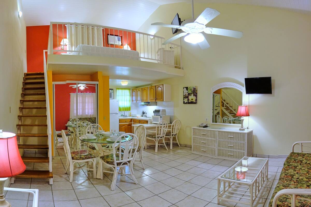 A spacious living area with a dining table, chairs, a sofa, a TV, and a staircase leading to an upper level.