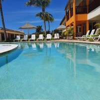 A clear swimming pool with lounge chairs and umbrellas, surrounded by palm trees and a building with balconies.