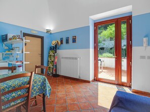 Ein Zimmer mit Esstisch, Stühlen, einem Heizkörper, einer Tür zu einem Außenbereich und einem Regal mit diversen Gegenständen.