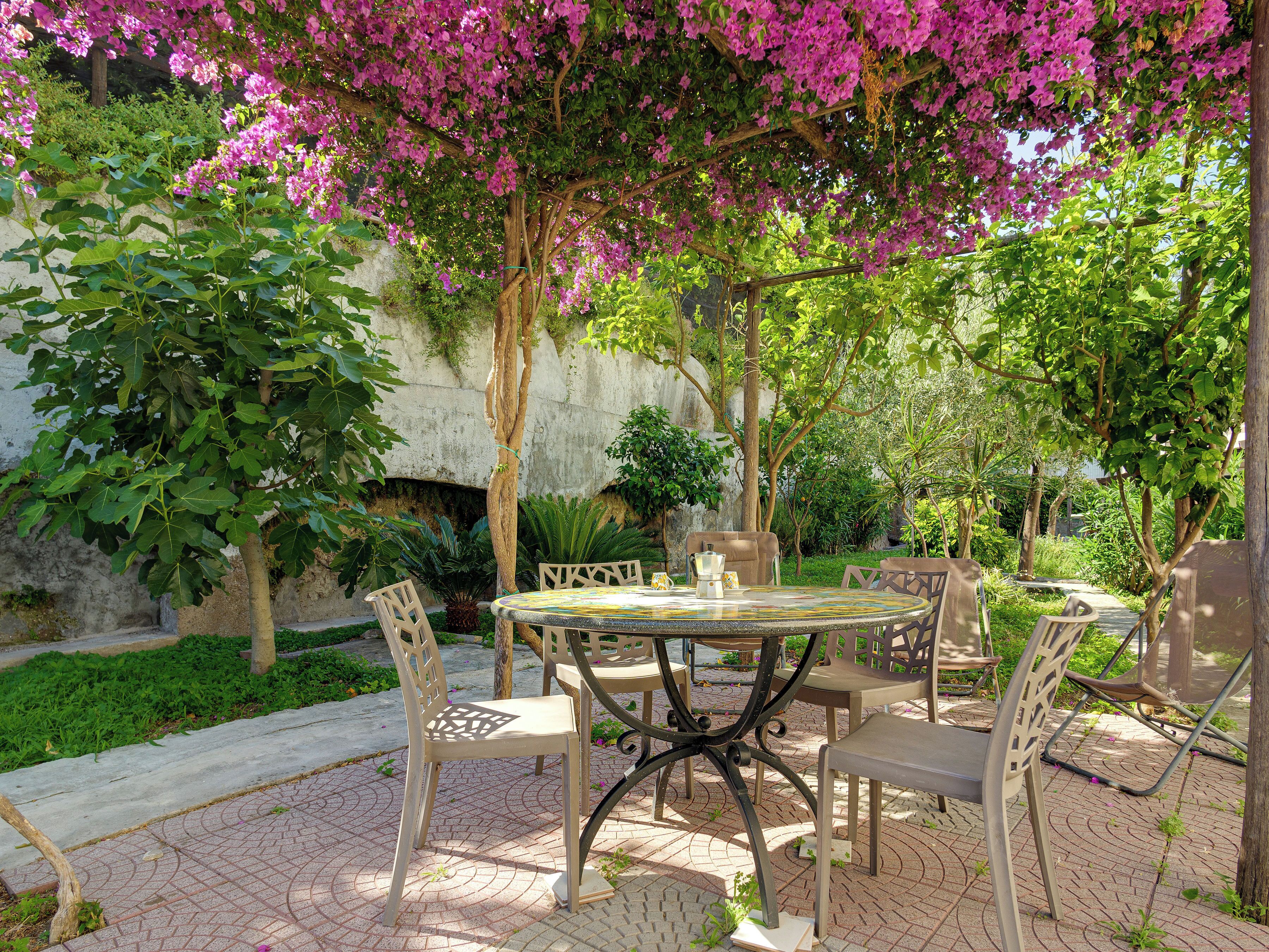 Eine Terrasse im Freien mit einem runden Tisch und Stühlen, umgeben von üppiger Bepflanzung und blühenden Bäumen.