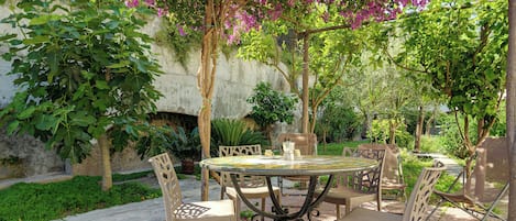 Un patio al aire libre con una mesa redonda y sillas, rodeado de abundante follaje y árboles en flor.