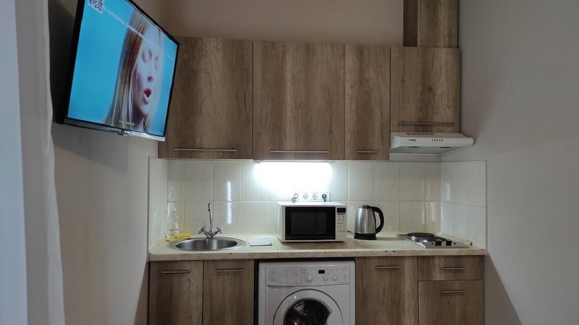A compact kitchen with wooden cabinets, a microwave, a kettle, a washing machine, and a sink.