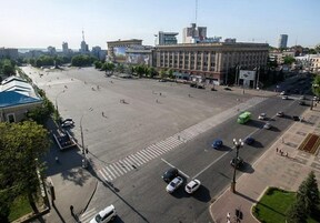 Vista aérea de una plaza urbana con edificios, algunos coches y cielo despejado.