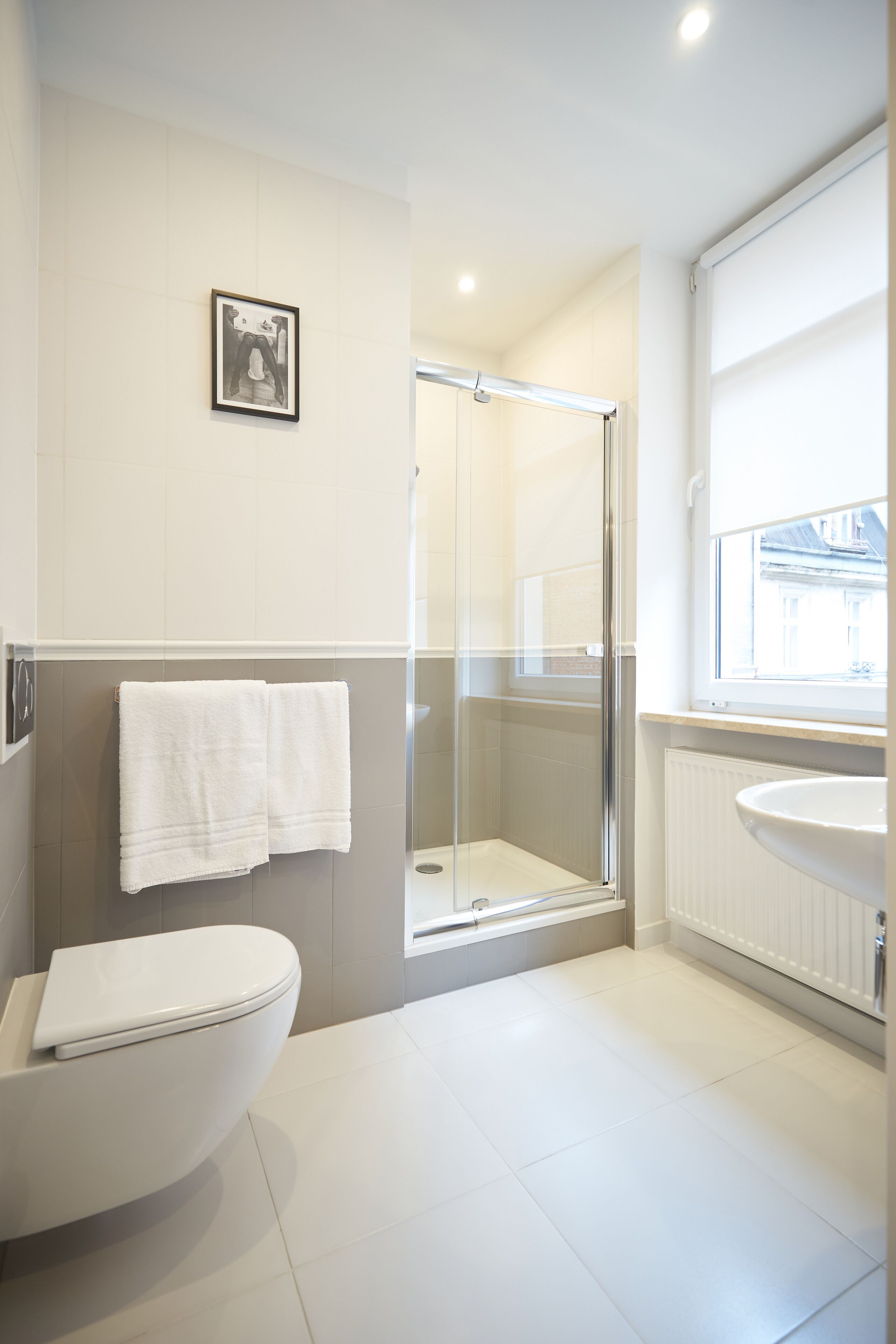 A modern bathroom with a white toilet, a glass shower enclosure, a white sink, and a window providing natural light.