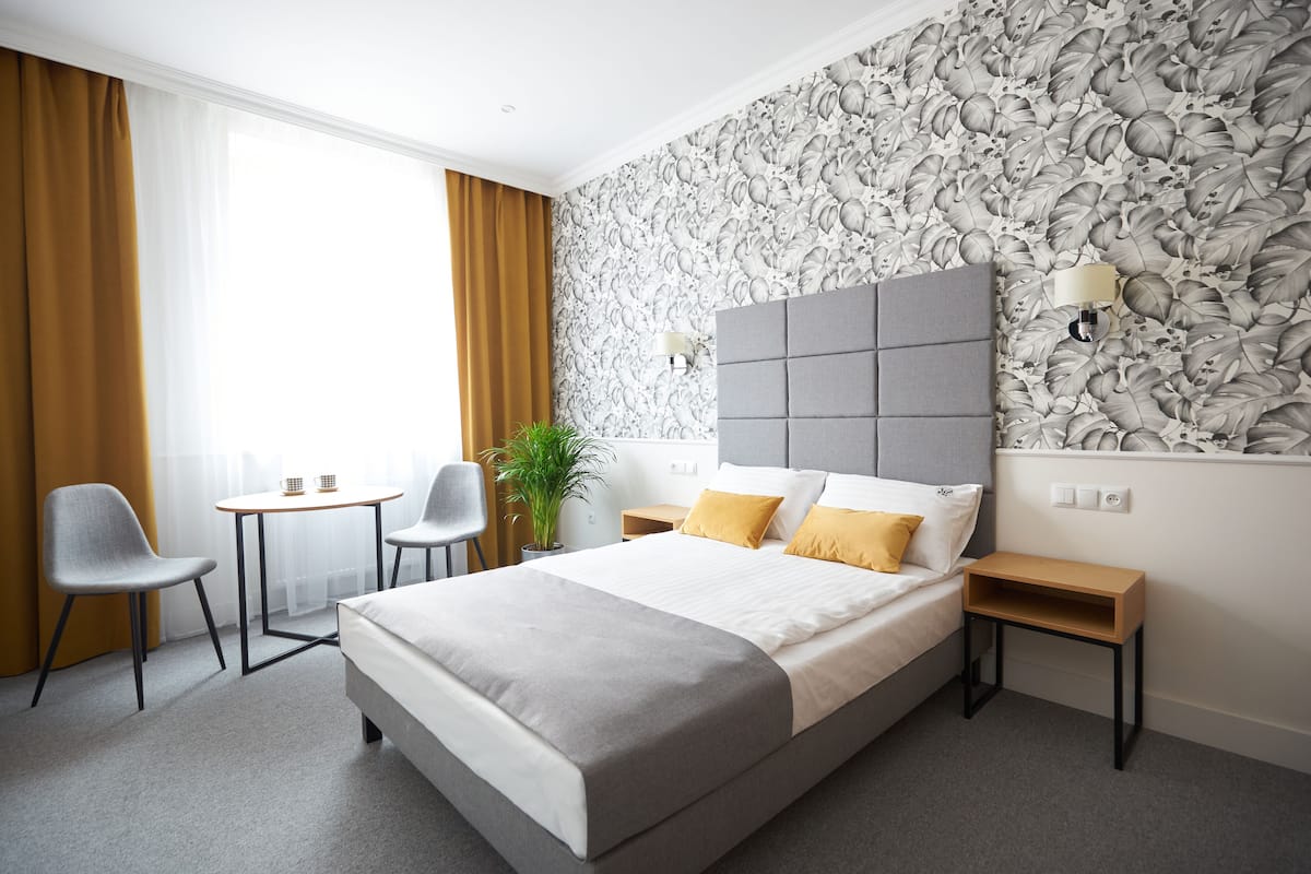 A modern hotel room with a large bed, a small table, two chairs, a potted plant, and a decorative wall.