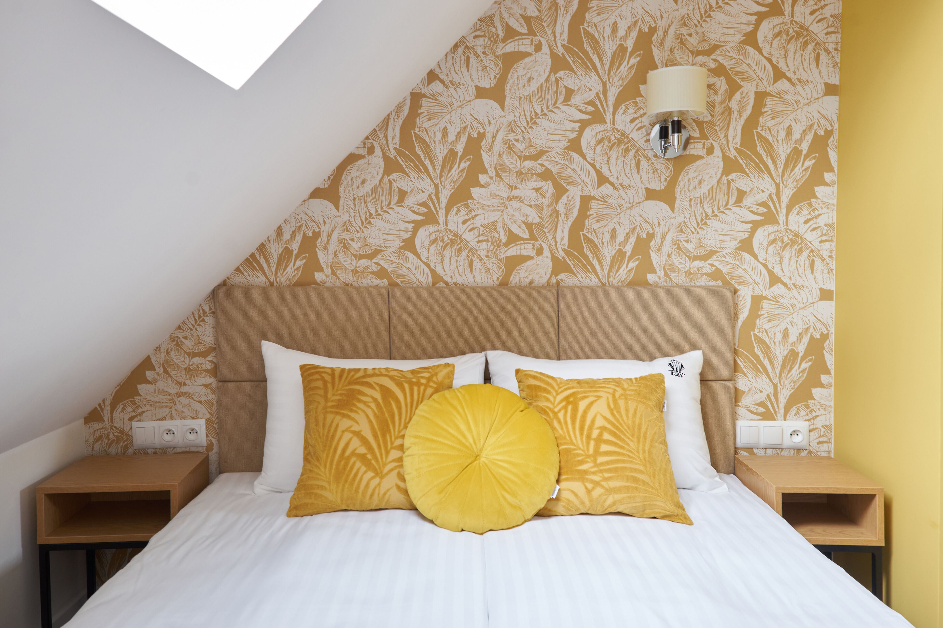 A bedroom with a sloped ceiling, a bed with white bedding and yellow pillows, a wall-mounted lamp, and a floral-patterned wallpaper.