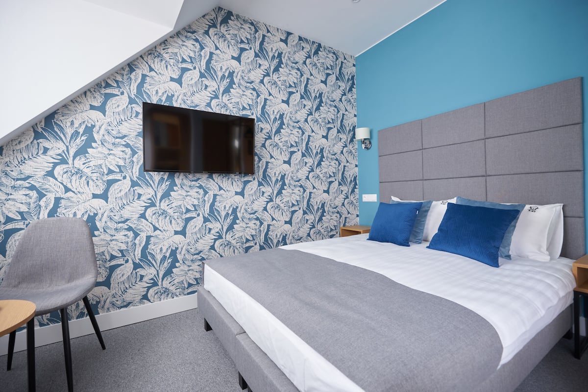 A modern hotel room with a large bed, a flat-screen TV mounted on the wall, a grey armchair, and a patterned wallpaper.