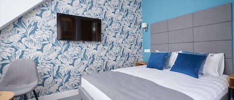 A modern hotel room with a large bed, a flat-screen TV mounted on the wall, a grey armchair, and a patterned wallpaper.