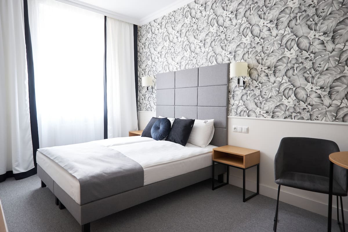 A modern hotel room with a large bed, a headboard with a light fixture, a small wooden nightstand, a desk, and a chair.