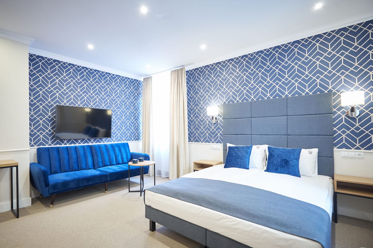 A modern hotel room with a large bed, a blue sofa, a flat-screen TV, and patterned walls.