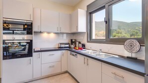 A modern kitchen with white cabinets, a built-in oven, and a window with a view of hills. - Nimfes Villas (Agios Nikolaos)