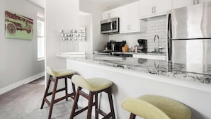 A modern kitchen with white cabinets, a stainless steel refrigerator, a microwave, and a granite countertop. There are three bar stools with cushioned seats.