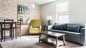 A living room with a brick wall, a yellow armchair, a grey sofa, a wooden coffee table, and a lamp. - Walk to Nightlife & Attractions (Indianapolis)