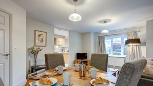A dining area with a wooden table set for breakfast, including croissants, coffee cups, and a teapot. There are four grey chairs around the table, and a television is visible in the background.