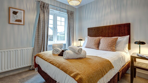 A neatly made bed with a brown throw blanket, a bedside table with a lamp, and a window with patterned curtains.
