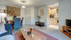 A modern living room with a sofa, dining table, and chairs. There is a kitchen area visible through an open door.