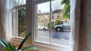 A view from a window with a white frame, looking out onto a street with parked cars and a brick building in the background. - Abbey Road | Oxford Holiday Let | 2 Beds & Sleeps 3 (Oxford)