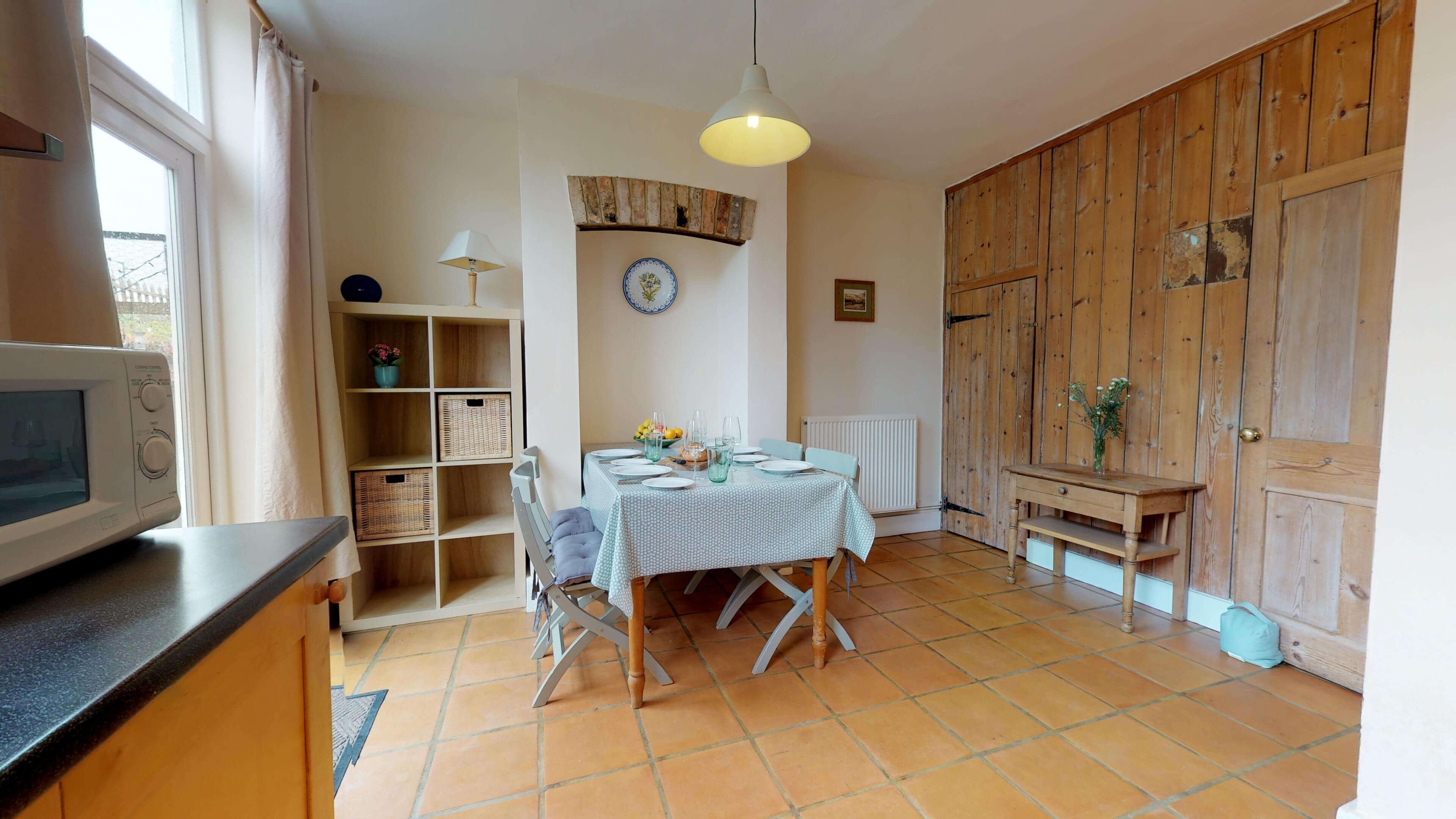 Una cocina con una mesa de comedor para dos personas, un microondas, una estantería con cestas y una puerta de madera.