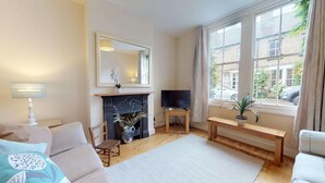 A living room with a fireplace, a television on a stand, a sofa, a chair, and a window with curtains. - Abbey Road | Oxford Holiday Let | 2 Beds & Sleeps 3 (Oxford)