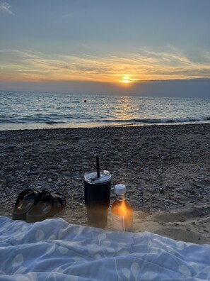 A beach scene with a sunset, a black thermos, a bottle, and a pair of sandals on the pebbly shore.